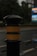 A close-up image of a black bollard with a yellow band, which is wet, likely due to rain. The background is blurred, showing a road, greenery, and possibly a sign or digital display board.