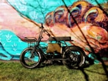 A black electric bicycle with thick tires is parked against a vibrant mural painted on a brick wall. The mural features abstract and colorful imagery, including organic shapes and claws in shades of teal, pink, orange, and blue. The scene is sunlit, casting tree branch shadows onto the wall and bicycle.