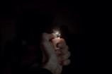 A hand is holding a lighter, which is producing a small flame and sparks. The background is dark, emphasizing the light and the details of the hand and lighter.
