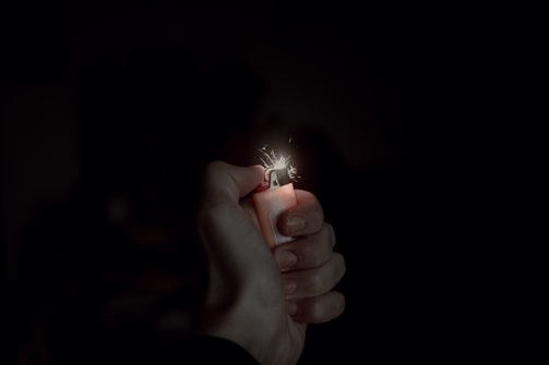 A hand is holding a lighter, which is producing a small flame and sparks. The background is dark, emphasizing the light and the details of the hand and lighter.