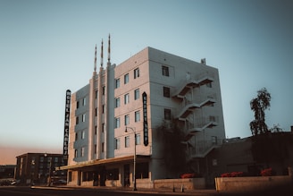 A multi-story building with a distinct Art Deco style, featuring vertical lines and decorative elements. The building facade is light-colored, with the words 'Hotel San Carlos' and 'Coffee Shop' visible on the signage. There is an exterior fire escape on the side of the building, and a streetlamp and a tree are present in the foreground.