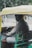A person is seated inside an auto rickshaw, holding the steering handle. The vehicle has a yellow roof and green body, with a meter device attached near the seat. The person is looking slightly towards the camera. In the background, there are green trees and an urban road scene.