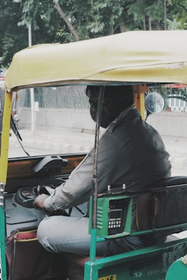 A person is seated inside an auto rickshaw, holding the steering handle. The vehicle has a yellow roof and green body, with a meter device attached near the seat. The person is looking slightly towards the camera. In the background, there are green trees and an urban road scene.