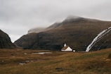 A small white church with a grass roof is nestled in a wide, grassy valley surrounded by steep, brown mountains. A thin waterfall cascades down the rocky slopes on the right side, and the sky is overcast with clouds hanging low over the mountains.