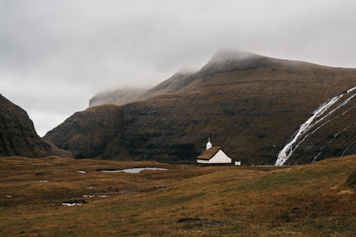 A small white church with a grass roof is nestled in a wide, grassy valley surrounded by steep, brown mountains. A thin waterfall cascades down the rocky slopes on the right side, and the sky is overcast with clouds hanging low over the mountains.