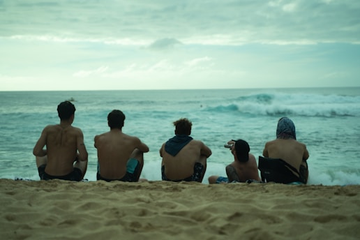 Five shirtless individuals sitting on a sandy beach facing the ocean. The waves are crashing in the background under an overcast sky.