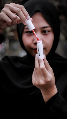 A person wearing a black hijab is holding a lip tint or lipstick. They are carefully removing the applicator from the tube, revealing a vivid red color. The focus is on the hands and the cosmetic product, while the background is blurred.