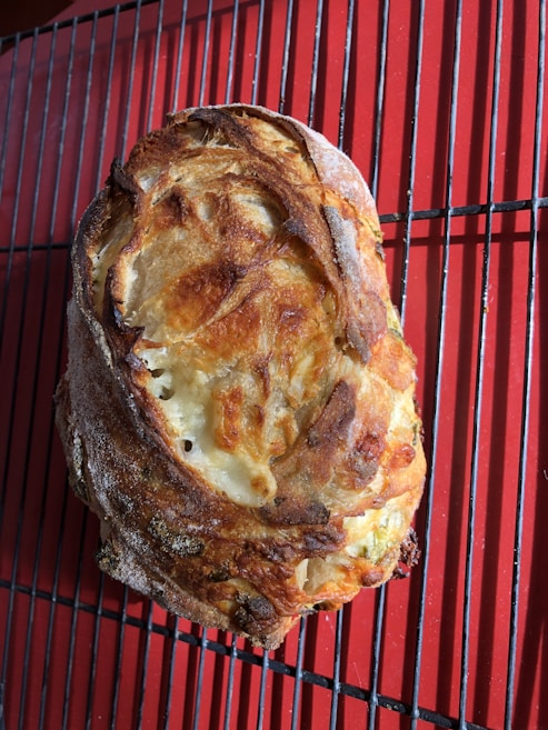 A freshly baked loaf of bread with a crispy, golden-brown crust rests on a red wire cooling rack. The bread's surface displays a variety of textures and colors, indicating a well-baked crust with some flour dusting.
