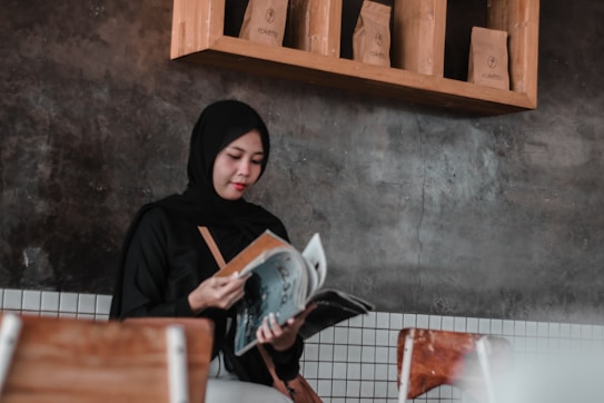 A person in a black hijab is sitting and reading a magazine in a room with a rustic gray wall and wooden shelving. The shelving holds brown paper bags labeled 'Raketna'. The seating consists of wooden chairs with white tiles visible on the lower part of the wall.