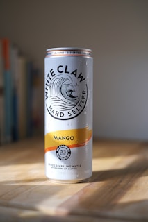 A can of White Claw Hard Seltzer Mango flavor, sitting on a wooden surface with a blurred background that appears to be a room with some books on a shelf. The can is tall and slender, predominantly silver with a yellow and orange stripe. The label highlights that it is gluten-free and has 100 calories.