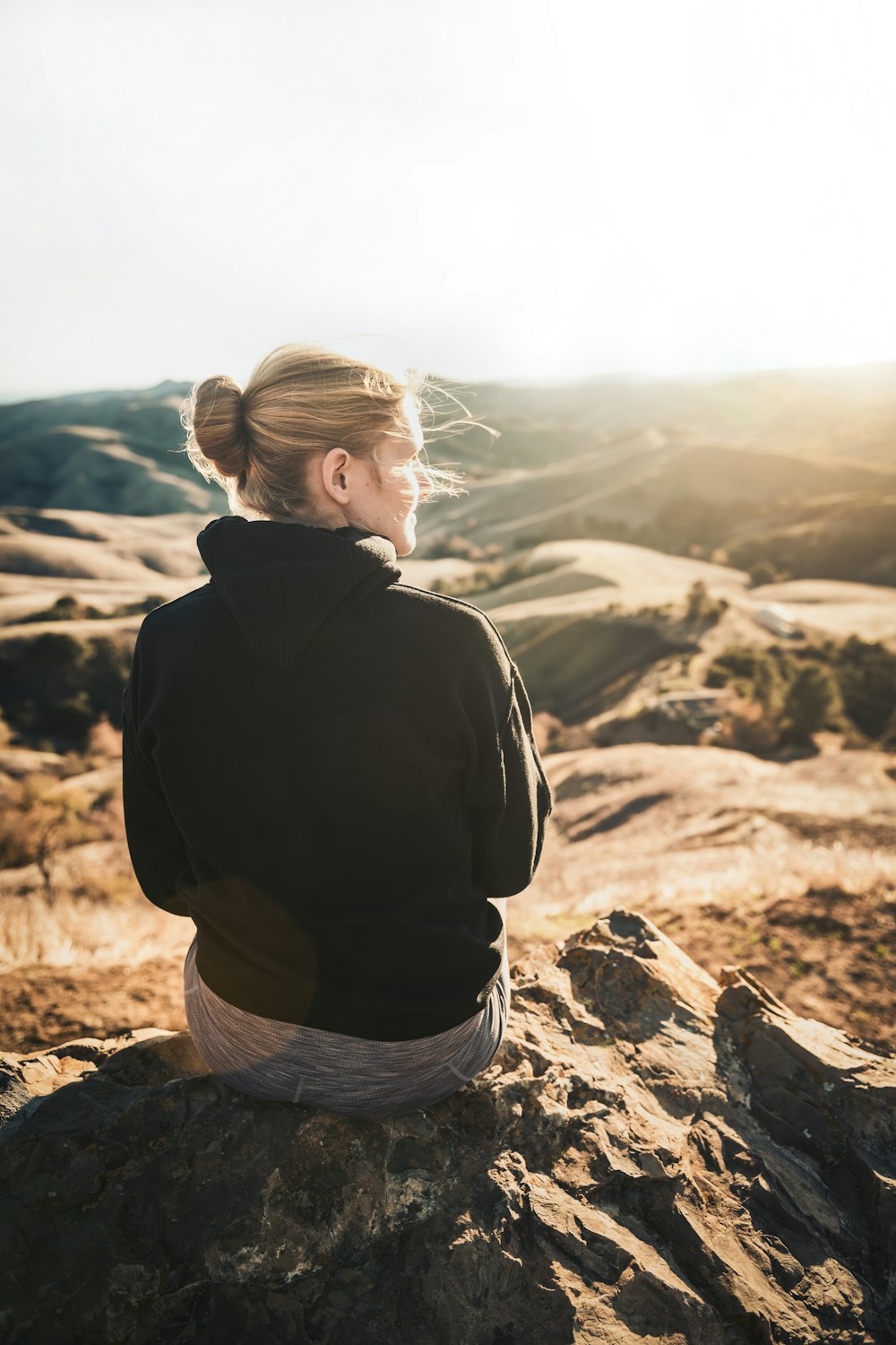 Mann in schwarzem Hoodie und grauen Jeansshorts sitzt tagsüber auf braunem Felsen