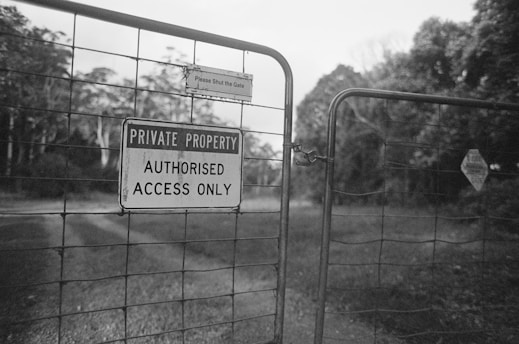 A metal gate with a sign that reads 'Private Property: Authorised Access Only' is closed. The gate is situated in a rural setting with trees and a dirt path visible in the background. Another smaller sign above requests individuals to 'Please Shut the Gate'.