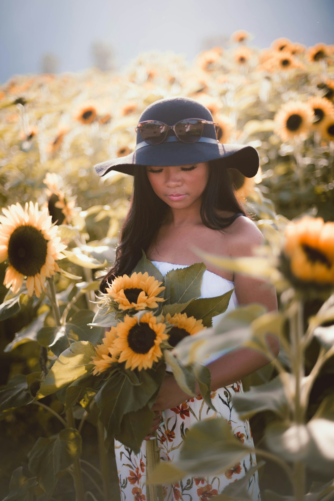 woman in black hat and white dress standing on sunflower field during daytime