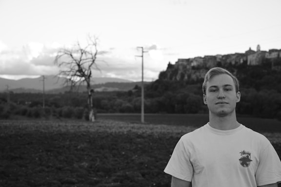 A black and white portrait of a young man standing in the foreground with a neutral expression. Behind him, a landscape features a leafless tree and a series of rolling hills, topped by a distant town or village. The sky is partly cloudy, adding to the serene and somewhat melancholic ambiance.