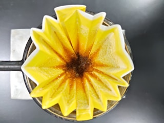 A top-down view of a coffee filter in a pour-over coffee maker. The yellow, star-shaped filter holds ground coffee, with the brewing process creating a rich brown color at the center.