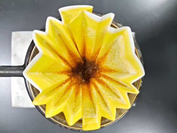 A top-down view of a coffee filter in a pour-over coffee maker. The yellow, star-shaped filter holds ground coffee, with the brewing process creating a rich brown color at the center.
