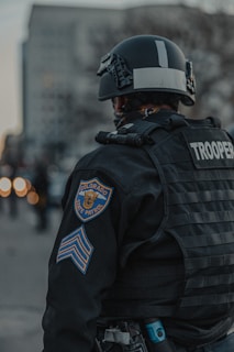 A Colorado State Patrol officer wearing tactical gear, including a helmet and body armor, is standing with their back facing outward. The officer is in an urban setting, with blurred city buildings visible in the background. The focus is on the uniform and patches indicating state trooper designation.
