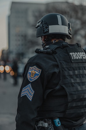 A Colorado State Patrol officer wearing tactical gear, including a helmet and body armor, is standing with their back facing outward. The officer is in an urban setting, with blurred city buildings visible in the background. The focus is on the uniform and patches indicating state trooper designation.