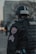 A Colorado State Patrol officer wearing tactical gear, including a helmet and body armor, is standing with their back facing outward. The officer is in an urban setting, with blurred city buildings visible in the background. The focus is on the uniform and patches indicating state trooper designation.