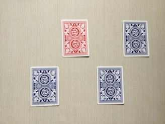 Four playing cards are arranged on a flat surface with a neutral background. Three of the cards have blue backs with intricate white patterns, and one card has a red back with a similar white pattern.