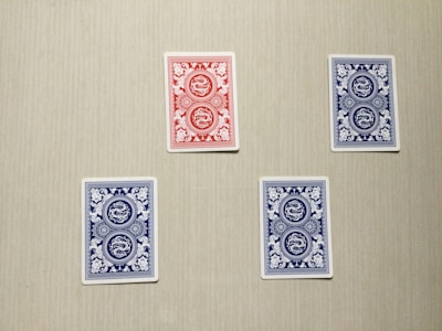 Four playing cards are arranged on a flat surface with a neutral background. Three of the cards have blue backs with intricate white patterns, and one card has a red back with a similar white pattern.