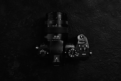 A black digital camera with a zoom lens is positioned against a dark textured background. The camera body features various control dials and buttons, including settings for ISO, white balance, and exposure compensation. The lens displays focal length markings, ranging from 12 to 35 mm.