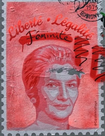 A stylized portrait of a woman is depicted on a textured surface resembling a stamp. The image is dominated by shades of red and pink. Written text in stylized fonts includes the words 'Liberté', 'Légalité', and 'Féminité'. The top right corner features a postmark stamp with the year 1975 and the location Bobigny. Black scribbles overlay parts of the image.
