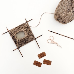 A small piece of knitting is in progress with four knitting needles positioned around it, accompanied by a ball of yarn. Nearby are a pair of ornate scissors and three small labels with sewing and knitting motifs.