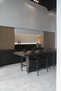 A modern kitchen features a long wooden island with several black bar stools surrounding it. The countertop is decorated with matching black bowls and plates. The background includes minimalistic cabinetry in a light wood tone, accented by under-cabinet lighting and decorative items like candleholders and a small plant.
