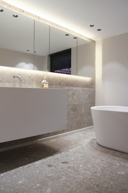 A minimalist bathroom features a sleek, modern design with a large white bathtub and a floating vanity with a smooth surface. Above the vanity, a set of mirrored cabinets extends across the wall, reflecting the light from recessed ceiling fixtures. The walls and floor are covered in light gray stone tiles, creating a cohesive and calming color scheme. The ambiance is enhanced by soft ambient lighting and a small diffuser on the vanity.