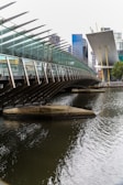 A modern bridge with a glass and steel structure stretches over a body of water. In the background, tall office buildings with a mix of glass and concrete rise into the skyline. The architectural style is contemporary, with sharp lines and angled surfaces.