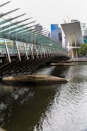 A modern bridge with a glass and steel structure stretches over a body of water. In the background, tall office buildings with a mix of glass and concrete rise into the skyline. The architectural style is contemporary, with sharp lines and angled surfaces.