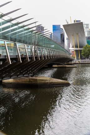 A modern bridge with a glass and steel structure stretches over a body of water. In the background, tall office buildings with a mix of glass and concrete rise into the skyline. The architectural style is contemporary, with sharp lines and angled surfaces.