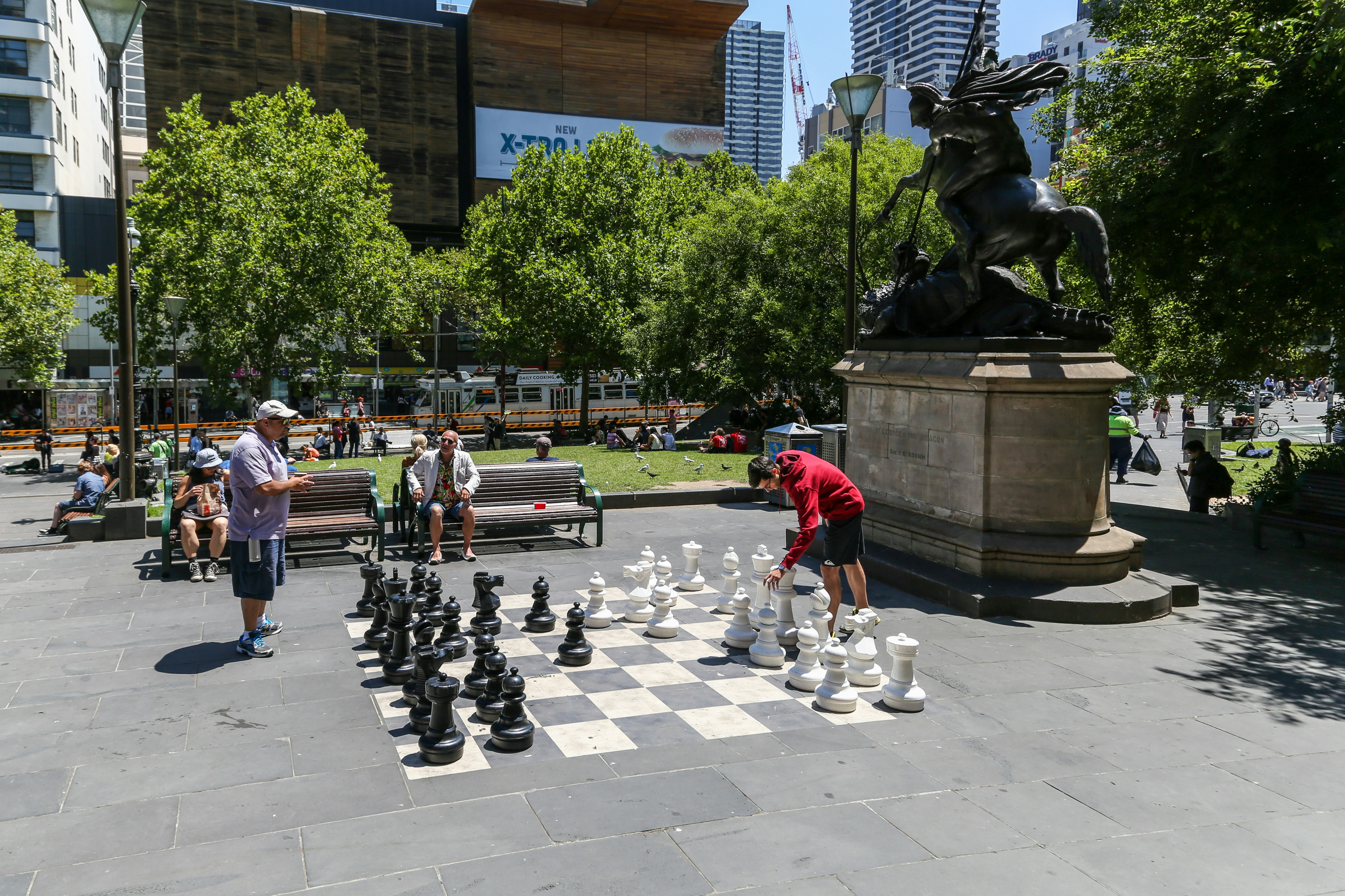 A public outdoor setting features a large chessboard with oversized black and white chess pieces. Two people are engaged in a game, watched by others seated on nearby benches. The scene includes a prominent statue mounted on a pedestal, with lush green trees and modern buildings in the background.