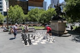 A public outdoor setting features a large chessboard with oversized black and white chess pieces. Two people are engaged in a game, watched by others seated on nearby benches. The scene includes a prominent statue mounted on a pedestal, with lush green trees and modern buildings in the background.