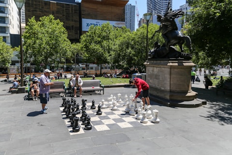 A public outdoor setting features a large chessboard with oversized black and white chess pieces. Two people are engaged in a game, watched by others seated on nearby benches. The scene includes a prominent statue mounted on a pedestal, with lush green trees and modern buildings in the background.