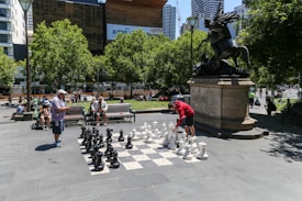 A public outdoor setting features a large chessboard with oversized black and white chess pieces. Two people are engaged in a game, watched by others seated on nearby benches. The scene includes a prominent statue mounted on a pedestal, with lush green trees and modern buildings in the background.