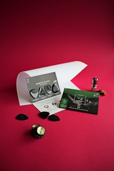 A collection of guitar picks and marketing material is displayed on a vibrant red surface. The picks come in various shapes, placed on or around two informational cards. One card is gray with images and descriptions of the picks, and the other is predominantly green. A smooth paper sheet partially rolls in the background. Additional elements include a small metallic tube and a gear-like object with a knob.