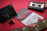 On a red background, there is a set of pink guitar picks with a diamond-like texture placed on top of a promotional card. Nearby is a vintage audio cassette tape along with some golden screw hardware. A decorative ribbon with intricate patterns accents the scene, suggesting a musical theme.