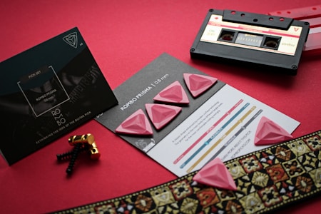On a red background, there is a set of pink guitar picks with a diamond-like texture placed on top of a promotional card. Nearby is a vintage audio cassette tape along with some golden screw hardware. A decorative ribbon with intricate patterns accents the scene, suggesting a musical theme.