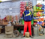 A street vendor stall displaying a variety of snack packets arranged in neat rows on the wall. A young person stands in front of the stall wearing a blue and yellow jacket and red pants, looking upward. The scene includes large metal pots and kettles on the counter, a small clay stove, and baskets containing cups.