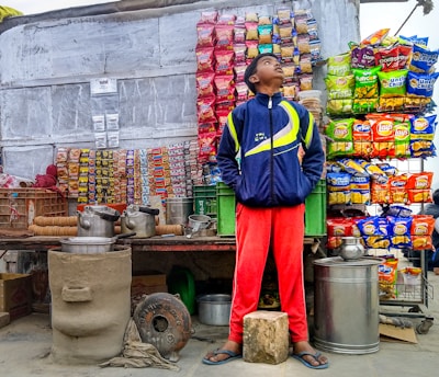 A street vendor stall displaying a variety of snack packets arranged in neat rows on the wall. A young person stands in front of the stall wearing a blue and yellow jacket and red pants, looking upward. The scene includes large metal pots and kettles on the counter, a small clay stove, and baskets containing cups.
