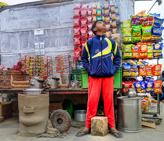 A street vendor stall displaying a variety of snack packets arranged in neat rows on the wall. A young person stands in front of the stall wearing a blue and yellow jacket and red pants, looking upward. The scene includes large metal pots and kettles on the counter, a small clay stove, and baskets containing cups.