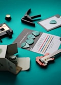 The image features a collection of music-related items arranged on a teal surface. There are several guitar picks on informational cards, a small clamp, a miniature plastic guitar model made from building blocks, and a small toy van partially inside a cardboard box. A cassette tape with a label is also visible.