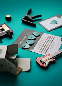 The image features a collection of music-related items arranged on a teal surface. There are several guitar picks on informational cards, a small clamp, a miniature plastic guitar model made from building blocks, and a small toy van partially inside a cardboard box. A cassette tape with a label is also visible.