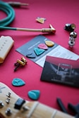 A collection of guitar-related items is spread out on a red surface. There are guitar picks of various colors and shapes, an electric guitar pickup, a green instrument cable, a vacuum tube, screws and hardware, and a guitar neck with an aqua-colored pick lying on it. Instruction cards and booklets are also visible.
