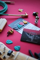 A collection of guitar-related items is spread out on a red surface. There are guitar picks of various colors and shapes, an electric guitar pickup, a green instrument cable, a vacuum tube, screws and hardware, and a guitar neck with an aqua-colored pick lying on it. Instruction cards and booklets are also visible.