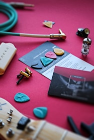 A collection of guitar-related items is spread out on a red surface. There are guitar picks of various colors and shapes, an electric guitar pickup, a green instrument cable, a vacuum tube, screws and hardware, and a guitar neck with an aqua-colored pick lying on it. Instruction cards and booklets are also visible.