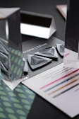 An arrangement of triangular and rectangular prisms sits on a dark background. The prisms are transparent and reflective, creating visually interesting reflections and refractions. Surrounding the prisms are pieces of paper with text and colored lines. A pattern of green and teal dots is visible on one sheet.