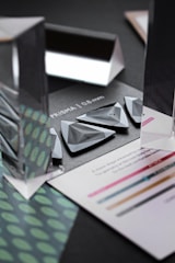 An arrangement of triangular and rectangular prisms sits on a dark background. The prisms are transparent and reflective, creating visually interesting reflections and refractions. Surrounding the prisms are pieces of paper with text and colored lines. A pattern of green and teal dots is visible on one sheet.